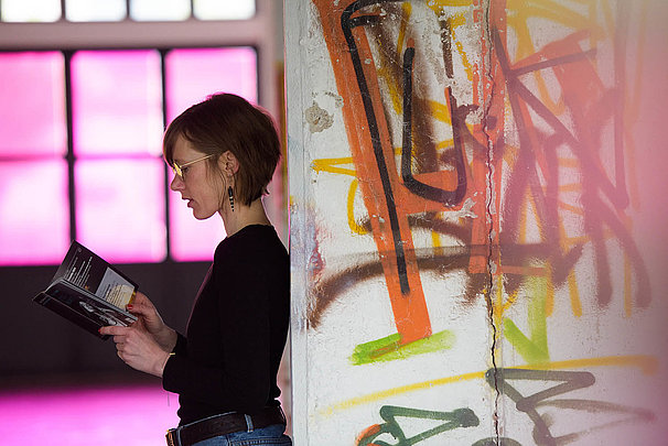  A woman leaning against a wall is reading a program of the Bach Festival