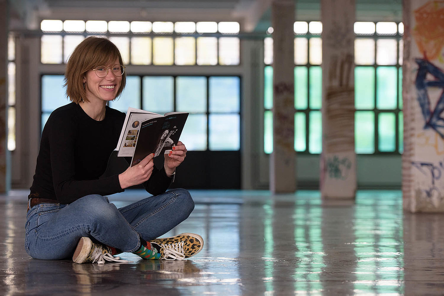 a woman sitting on the floor holds a program of the Bach Festival in her hands and smiles friendly at the camera