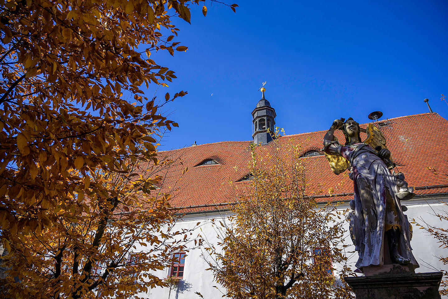 Im Vordergrund steht eine Steinskulptur eines Engels mit goldenen Flügeln. Im Hintergrund das Rathaus von Buttstädt