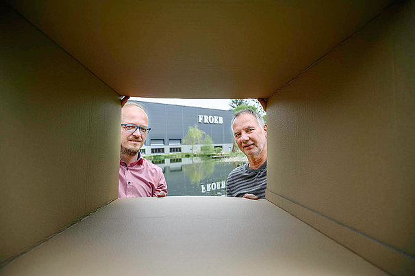 Two men look at the camera through a cardboard box. Behind them is a house belonging to Froeb-Ver Verpackungen GmbH.