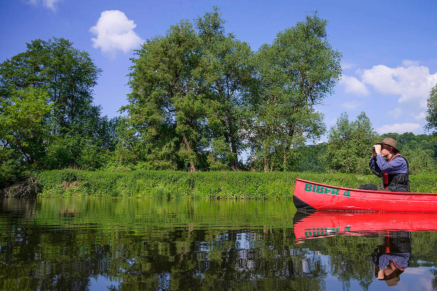 ein durch ein Fernglas schauender Mann fährt mit einem Kanu über einen Fluss