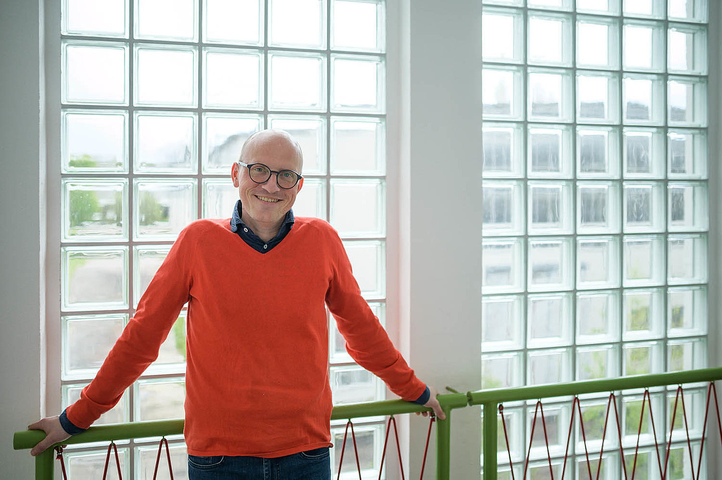 a man in an orange sweater leans against a railing and smiles friendly at the camera