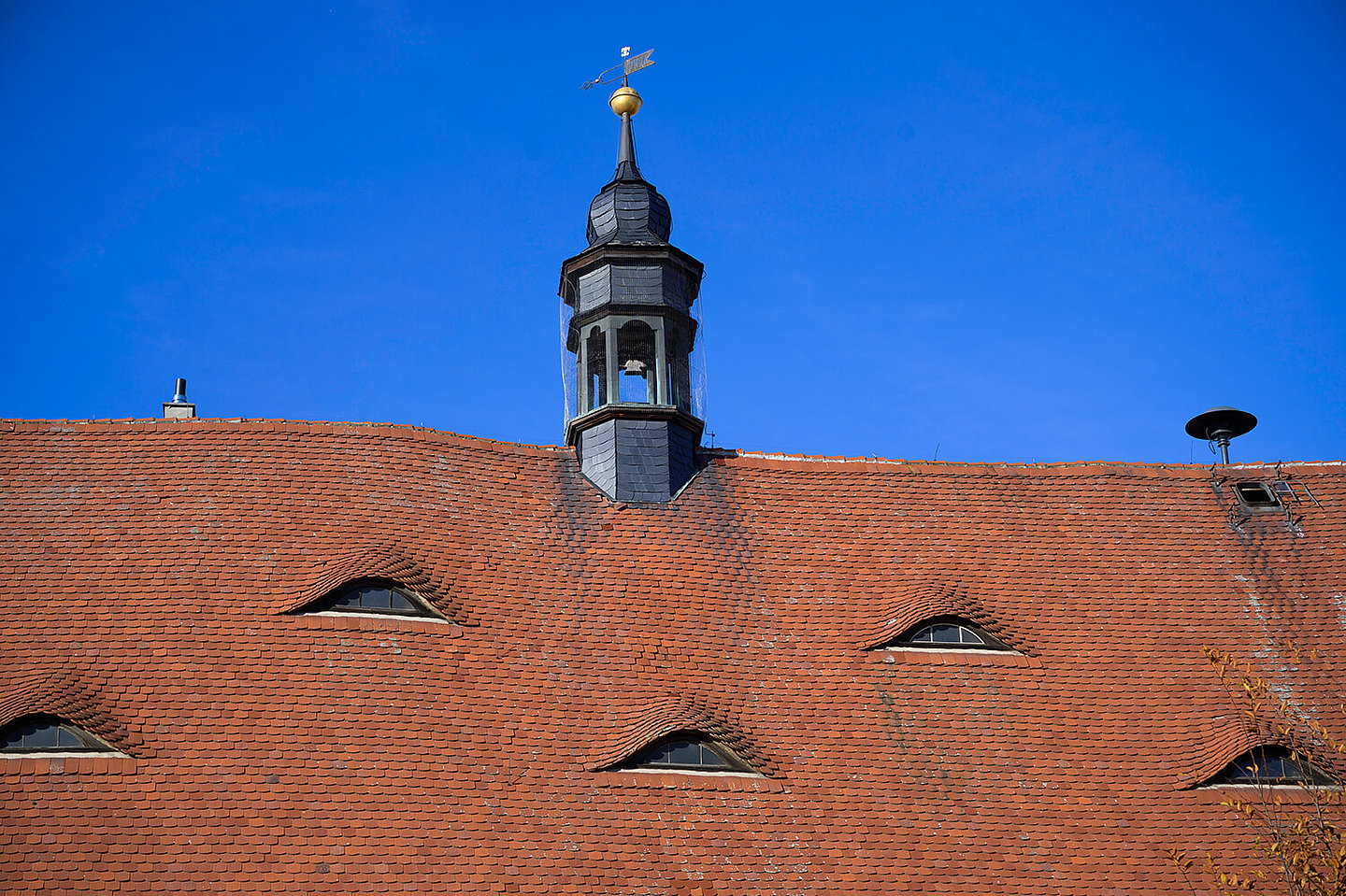 Glockenturm vom Buttstädter Rathaus. Dieser sitzt auf einem roten Dach mit kleinen Fenstern. In einer Ecke ist eine Alarmsirene montiert.